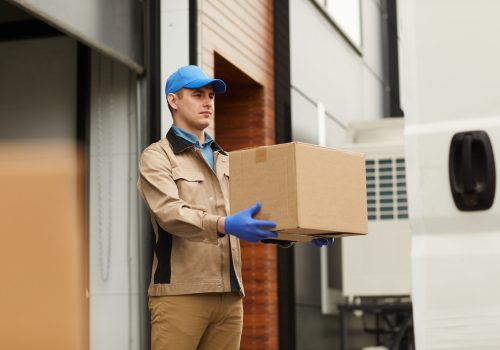 Young courier in uniform carrying cardboard box to the delivery van